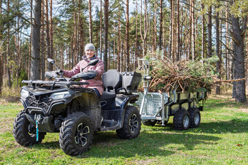 Fototapeta premium A woman drives a black all-terrain vehicle pulling a silver trailer packed with tree branches along a forest track. She wears a pink jacket and grey clothing while navigating.
