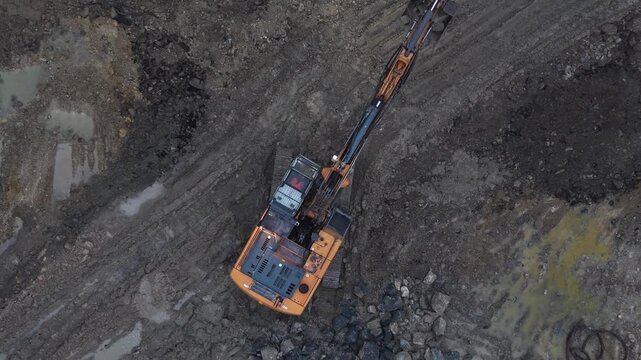 Excavator working with rocks and mud on construction site. Aerial top-down ascending