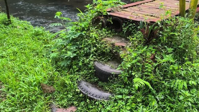 Rustic Outdoor Staircase Made of Recycled Rubber Tires Covered in Lush Greenery Leading to a Traditional Bamboo Bridge Over a Small River in a Tropical Village Environment