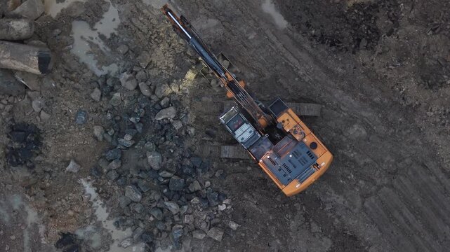 Excavator with lights on, working with rocks and mud on construction site at dusk. Aerial top-down view