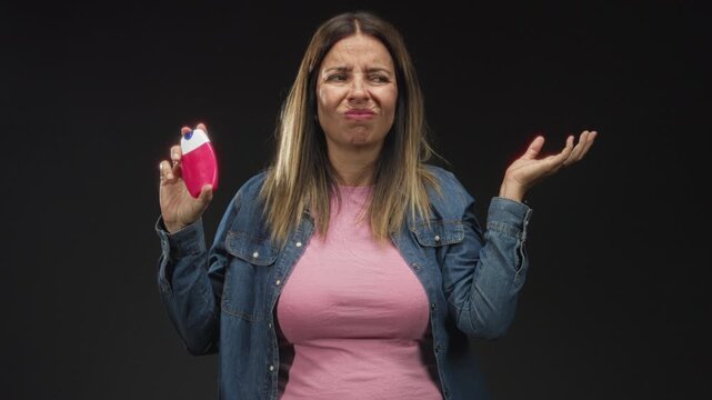 Woman holds a pink toothpaste dispenser in one hand, touches her head with the other hand and grimaces while wearing a denim jacket and pink shirt in a studio; confusion hesitation.