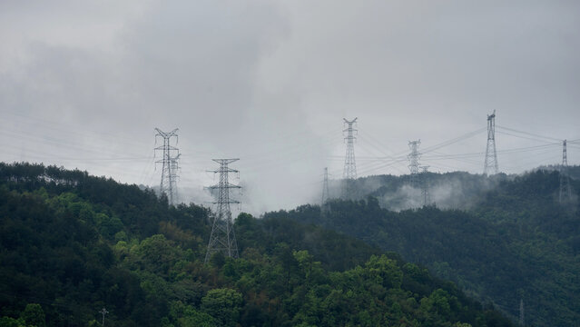 High-voltage transmission towers stand amidst misty mountains with dense forest and overhead power lines