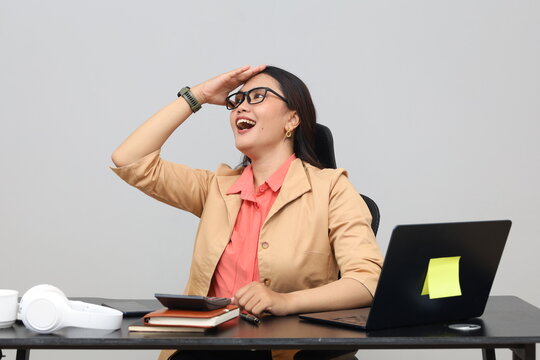 A young woman sits working at work with a laptop, a gesture of slapping her forehead while looking up, a facial expression of forgetting something.