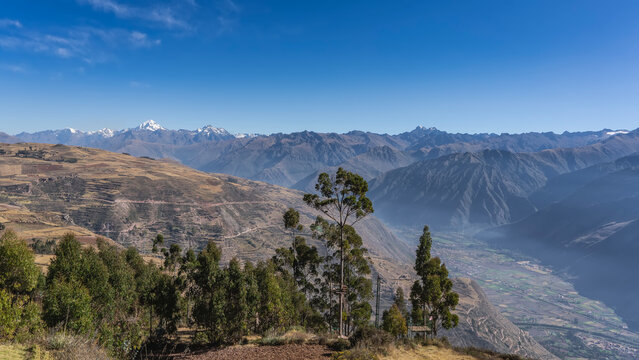 The landscape of the highlands. There are terraces on the mountain slopes, cultivated agricultural fields, green trees. There is a mountain range on the horizon. Snow-capped peaks, the blue sky. Peru
