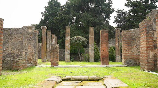Wide view of a Pompeii courtyard with standing columns, brick pillars, and a central stone feature, capturing the open garden layout of the ancient Roman house ruins.