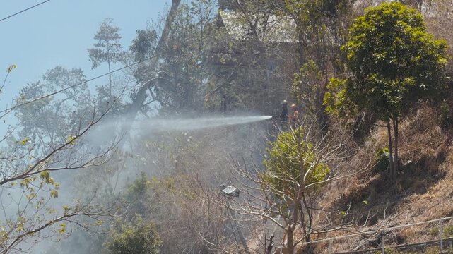 Firefighters spray water on burning hillside vegetation with smoke rising, static camera angle. &middot; 4K DCI 25fps