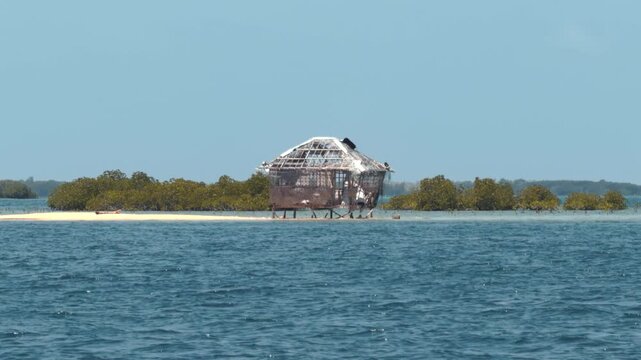 Static shot of a deteriorating structure on stilts surrounded by turquoise water, mangroves, and sandy shores in a tropical setting. &middot; 4K DCI 25fps