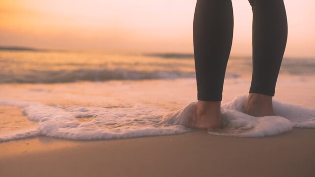 Close-up of legs in black athletic tights standing in sea foam with gentle waves splashing around feet at dusk. Perfect for wellness and healthy lifestyle content.