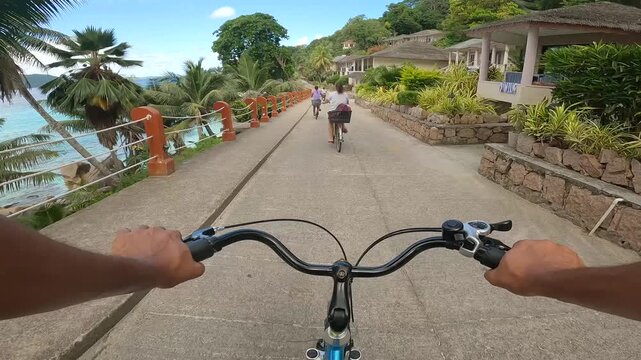 POV shot of family riding bicycles on a scenic paved path next to a beautiful tropical island beach.