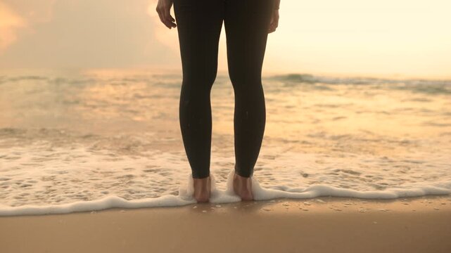 Close-up of legs in black athletic tights standing in sea foam with gentle waves splashing around feet at dusk. Perfect for wellness and healthy lifestyle content.