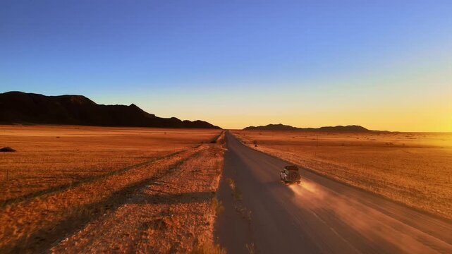 A jeep drives along a dusty road through the Namib Desert at sunset, filmed by drone, surrounded by glowing red dunes and golden light, capturing the wild beauty and freedom of desert adventure.