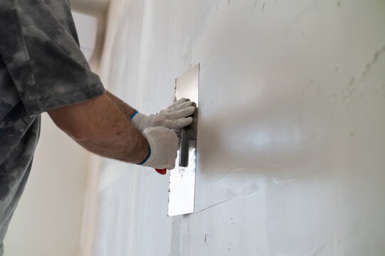 Skilled technician smoothing finishing compound with a stainless steel trowel on drywall during home renovation. Clean workspace environment with partially finished walls visible in the background.