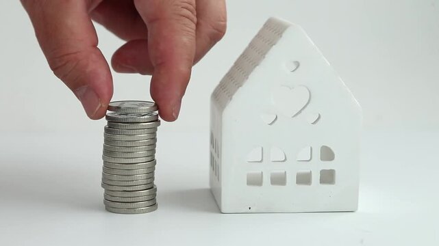 Male hand carefully stacks and builds a neat vertical column of silver coins placed right next to small white ceramic house figurine on white surface as savings concept