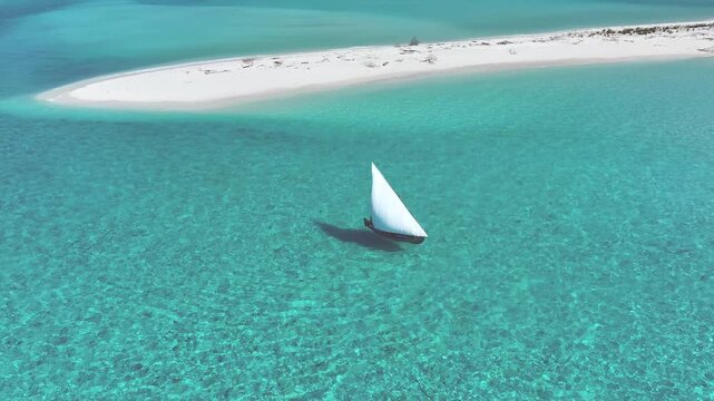 Aerial view of a traditional dhoni sailboat with a white sail gliding through turquoise waters near a pristine sandbank, Bileffahi, Shaviyani Atoll, Maldives.