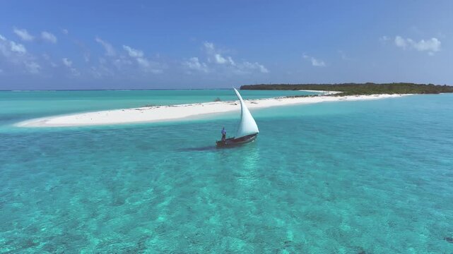 Aerial view of a traditional dhoni sailboat with a white sail gliding through turquoise waters near a white sand beach and lush tropical island in Bileffahi, Shaviyani Atoll, Maldives.