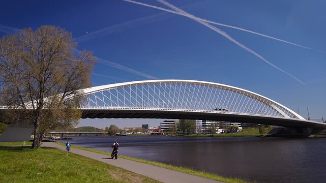 Low angle view of the modern white Troja Bridge in Prague spanning the Vltava river with a cyclist on a riverside path under a sunny blue sky
