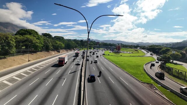 Elevated view of a multi-lane expressway with flowing cars and motorcycles surrounded by green trees and distant mountains in Venezuela.