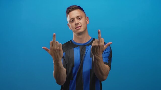 Young blond man in blue and black soccer jersey shows both middle fingers in a studio setting; defiance attitude.