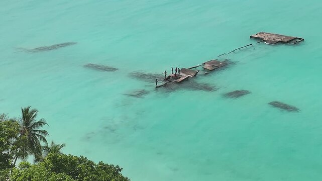 Aerial view of people standing on a broken wooden pier surrounded by turquoise tropical water and palm trees in Bileffahi, Shaviyani Atoll, Maldives.
