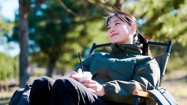 Slow motion tracking shot of young woman sipping drink and relaxing in park chair under pine trees on sunny day