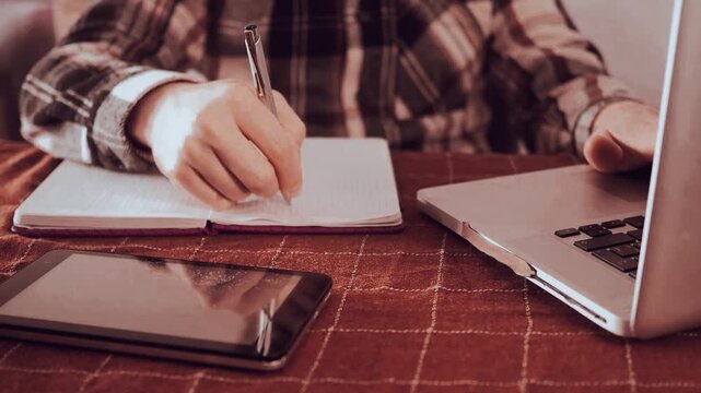 Close-up hands of male student writing notes with pen on notebook, reading book sitting at table in dark home setting.