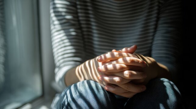 A person sits near a window, sunlight casts shadows, creating a thoughtful atmosphere. The person's hands are clasped, suggesting introspection and self-reflection