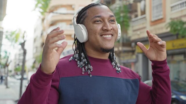 Man with headphones touching earcup and raising hands, smiling with beaded braids and maroon sweatshirt on a busy city street; joyful relaxation groove.