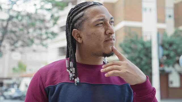 Man with braided hair and beaded ends, wearing burgundy sweater, finger to chin gesture on street; thoughtful reflection.