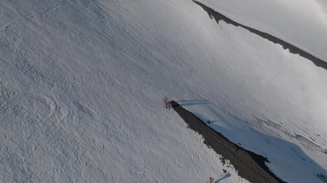 Aerial view of people standing on a volcanic patch amidst the snowy slopes of Mount Etna near Rifugio Sapienza in Nicolosi, Sicilia, Italy.