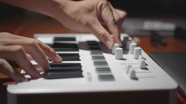 Close-up of human hands playing keys and adjusting knobs on a white compact MIDI keyboard controller in a music studio.