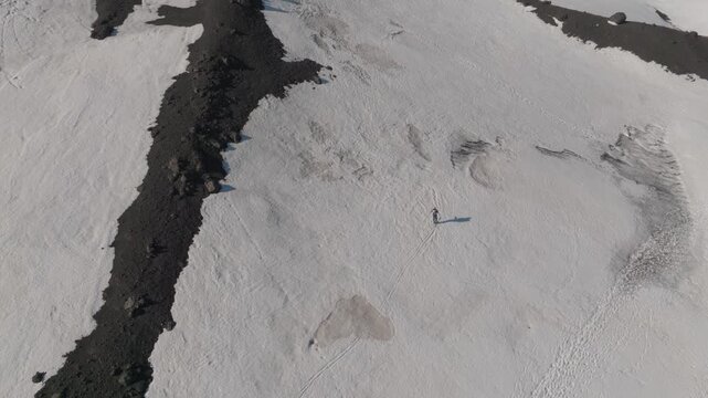 Aerial view of a lone hiker walking across a vast snowfield next to a dark volcanic ridge near Rifugio Sapienza in Nicolosi, Sicilia, Italy.