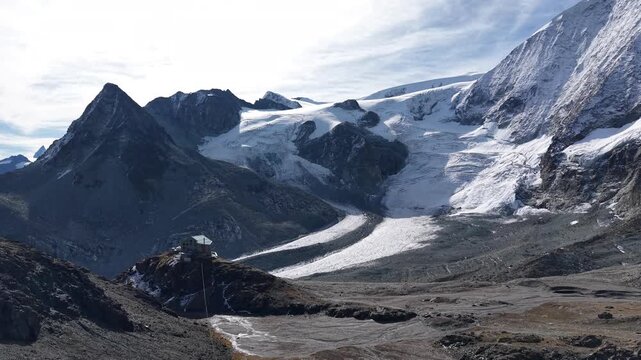 Aerial flyover of a mountain hut alongside Glacier de Cheilon as the morning sun shines over the mountain peaks of Swiss Alps in the Arolla region