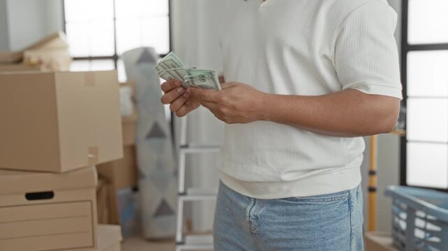 Man counting bills with hands beside stacked moving boxes in building, wearing white shirt and jeans; confidence savings.