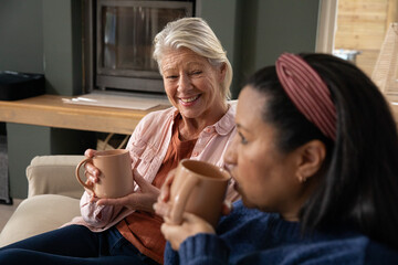 Naklejka premium Senior and mature female friends smiling and sipping tan mugs on beige sofa in living room