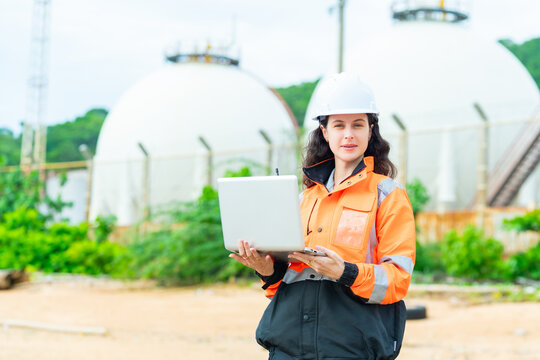 Female petroleum engineer using laptop computer at oil storage tank facility, energy industry worker wearing safety helmet and jacket working at industrial refinery fuel terminal operation site