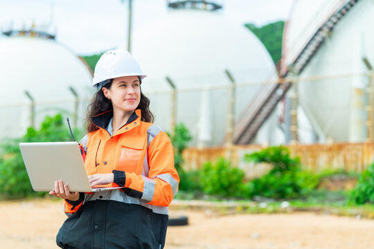 Female petroleum engineer using laptop computer at oil storage tank facility, energy industry worker wearing safety helmet and jacket working at industrial refinery fuel terminal operation site