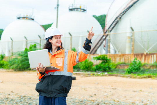 Female petroleum engineer using laptop computer at oil storage tank facility, energy industry worker wearing safety helmet and jacket working at industrial refinery fuel terminal operation site