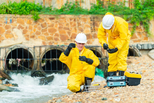 Environmental scientists collecting water samples near industrial drainage system, hazardous material inspectors in protective suits performing wastewater pollution testing environmental monitoring