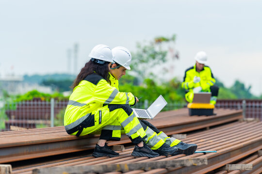 Industrial engineers using laptop while inspecting steel materials at construction site, technician wearing safety uniforms and helmets performing engineering analysis and infrastructure management