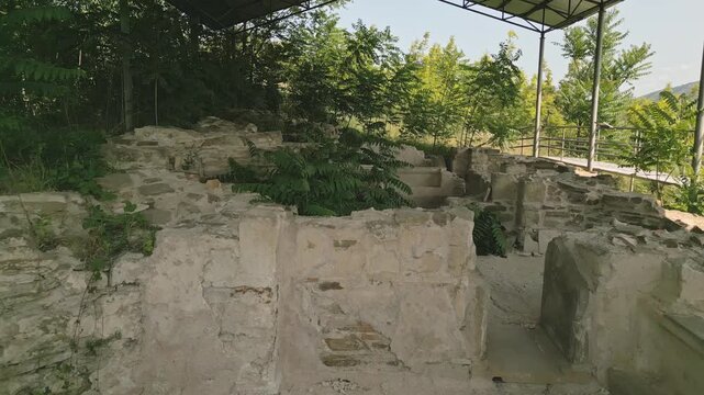 Walking point of view through Preserved remains of the ancient royal bath structure at Armaziskhevi site, surrounded by greenery under protective roof in Mtskheta region, Georgia