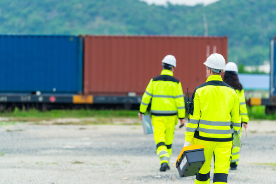 Industrial engineers walking at cargo container yard carrying equipment and laptop computers. Logistics workers wearing safety uniforms and helmets at transportation and shipping terminal site