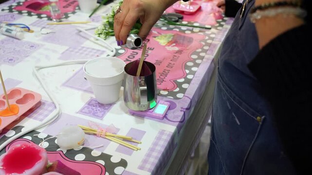 Artisan carefully pouring liquid scent into a metal pitcher with melted wax on a digital scale