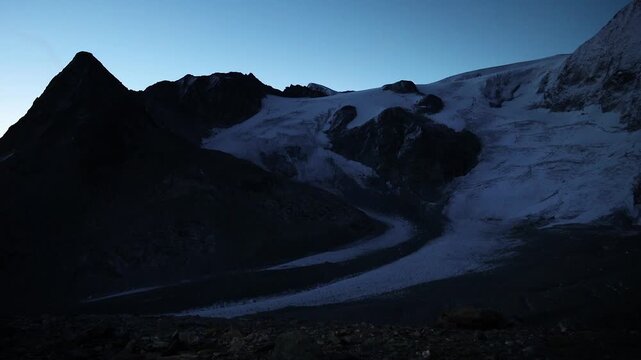 A view of a hiker overlooking Glacier de Cheilon during a sunrise hike with a shining headlamp with the mountain peaks of the Swiss Alps in the background