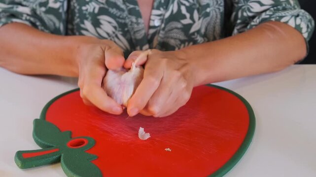 Woman peeling garlic cloves with knife, front view of removing thin skin before cooking, hands preparing fresh ingredient on cutting board, food preparation process in home kitchen