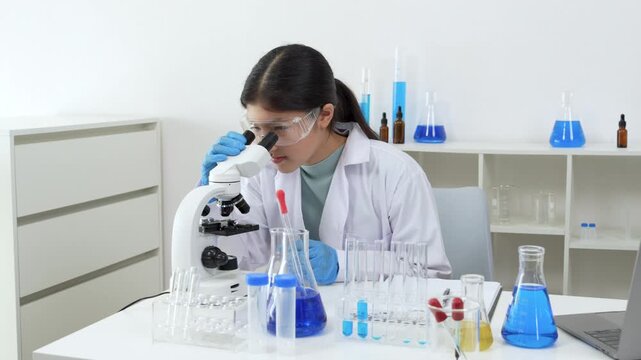 Young Asian women scientist working with test tube in laboratory. Scientist in a lab coat and safety glasses handles a test tube at a desk in a laboratory, make note on laptop, experimentation in a co