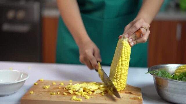 Close up of hands cutting corn kernels from the cob with a knife on a wooden cutting board