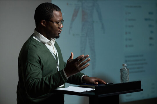 African American middle aged man presenting medical data beside anatomy screen, speaking at podium during seminar. Useful for healthcare training, conference promotion, expert education