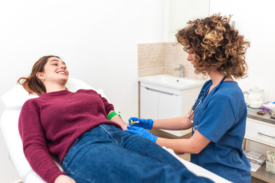 Female Doctor Drawing Patient Blood. Medical phlebotomy and healthcare blood test concept. Hospital And Clinic setting. Doctor with blue gloves inserting IV needle into smiling young woman's arm.
