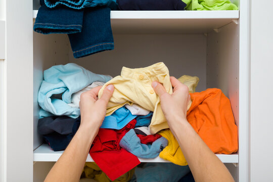Mother hands sorting cluttered different colorful clothes on shelves in white opened wooden wardrobe at home room. Messy casual kid apparel. Making neatness. Closeup. Point of view shot. Front view.