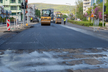 Fototapeta premium Two road rollers work in tandem to flatten a wide stretch of fresh, dark asphalt on a city boulevard during an extensive road renovation project.